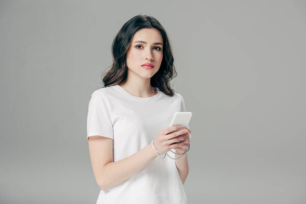 attractive young woman in white t-shirt and usb cable around hands using smartphone isolated on grey