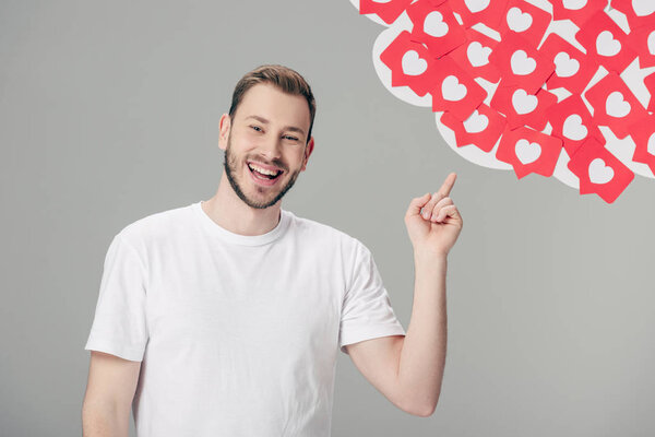 cheerful young man in white t-shirt pointing with finger at red paper cut cards with hearts symbols isolated on grey