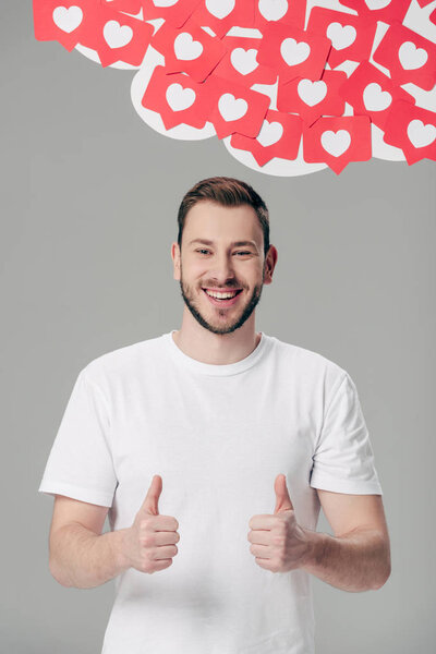 handsome young man in white t-shirt showing thumbs up near red paper cut cards with hearts symbols isolated on grey