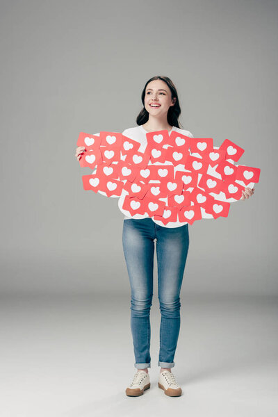 beautiful smiling girl in blue jeans holding red paper cut cards with hearts symbol on grey background