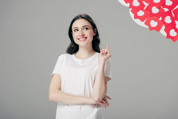 pretty young girl showing idea sign and looking up near red paper cut cards with hearts symbol isolated on grey