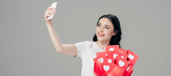panoramic shot of smiling girl holding bouquet of red paper cut cards with hearts symbol and taking selfie with smartphone isolated on grey