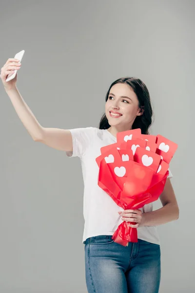 心のシンボルと赤い紙カットカードの花束を持ち 灰色で隔離されたスマートフォンで自分撮りを取る陽気な女の子 — ストック写真