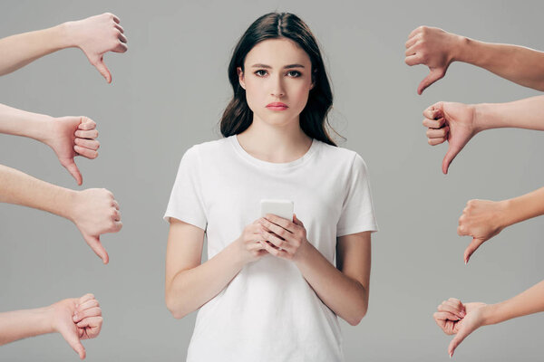 обрезанный вид мужчин и женщин, показывающих большие пальцы вниз около расстроенной девушки со смартфоном изолированы на серый
