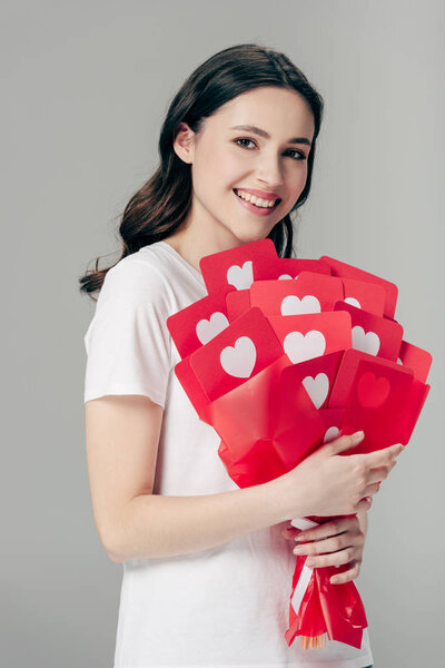 happy pretty girl holding bouquet of red paper cut cards with hearts symbols and looking at camera isolated on grey
