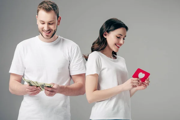 灰色で隔離されたドル紙幣を数えるハンサムな男の近くにハートのシンボルを持つ赤い紙カットカードを持つ魅力的な女の子 — ストック写真