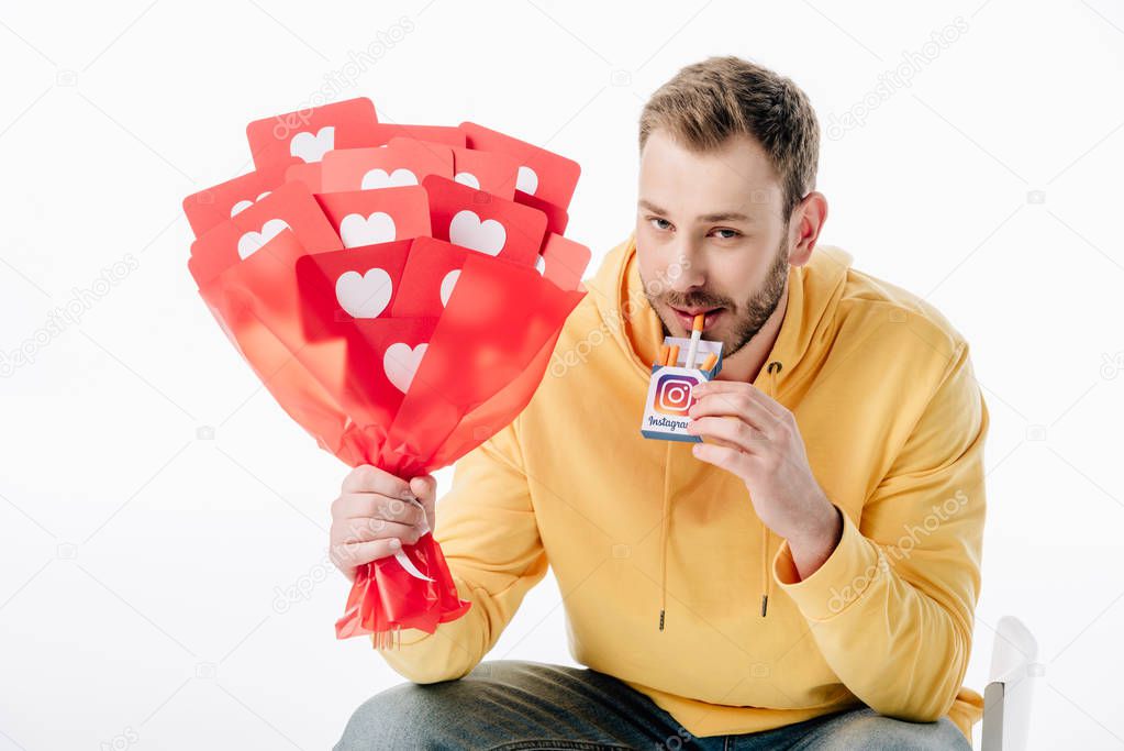 Handsome man holding cigarette pack with instagram logo and bouquet of red paper cut cards with hearts symbols isolated on white