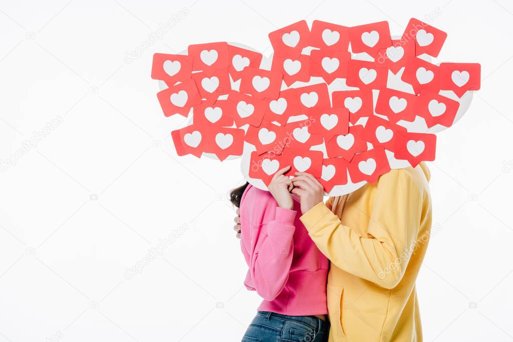 young man and woman in bright hoodies hiding behind faces behind red paper cut cards with heart symbols isolated on white