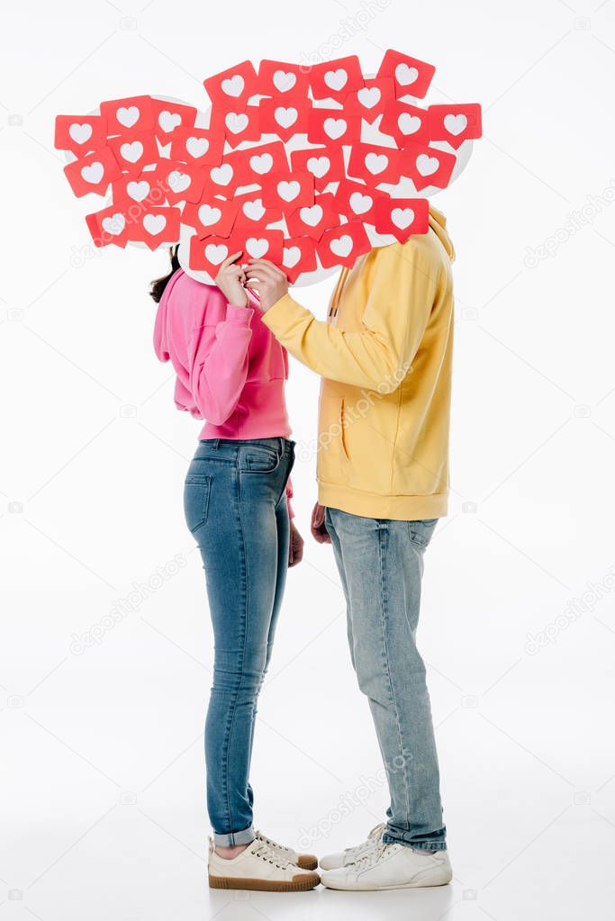 young couple in blue jeans and hoodies hiding faces behind red paper cut cards with hearts symbols on white background 