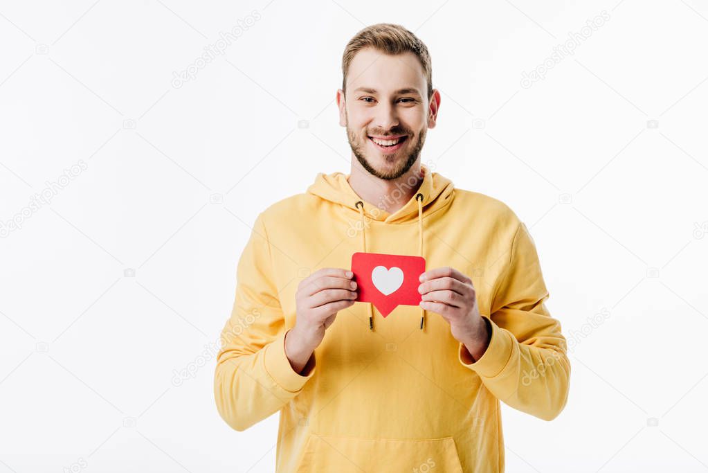 handsome cheerful man in yellow hoodie holding red paper cut card with heart symbol isolated on white