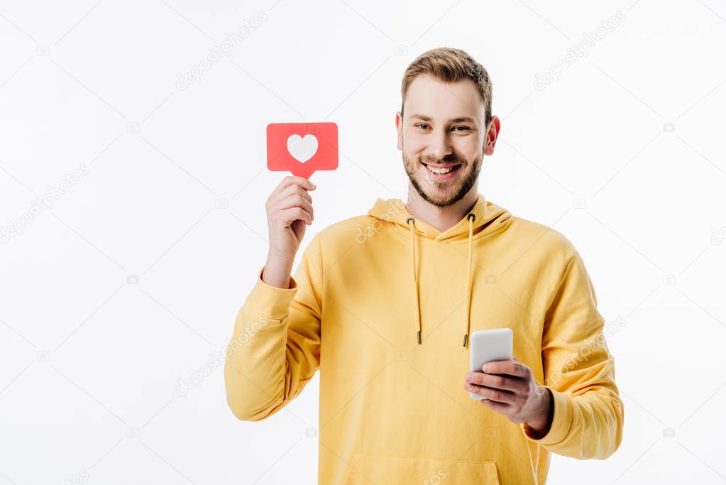 cheerful young man in yellow hoodie holding red paper cut card with heart symbol while using smartphone isolated on white