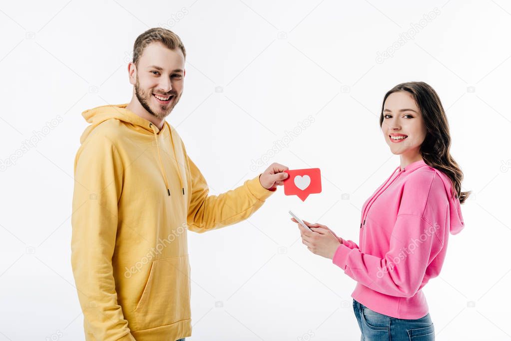 smiling handsome man giving red paper cut card with heart symbol to pretty girl using smartphone isolated on white