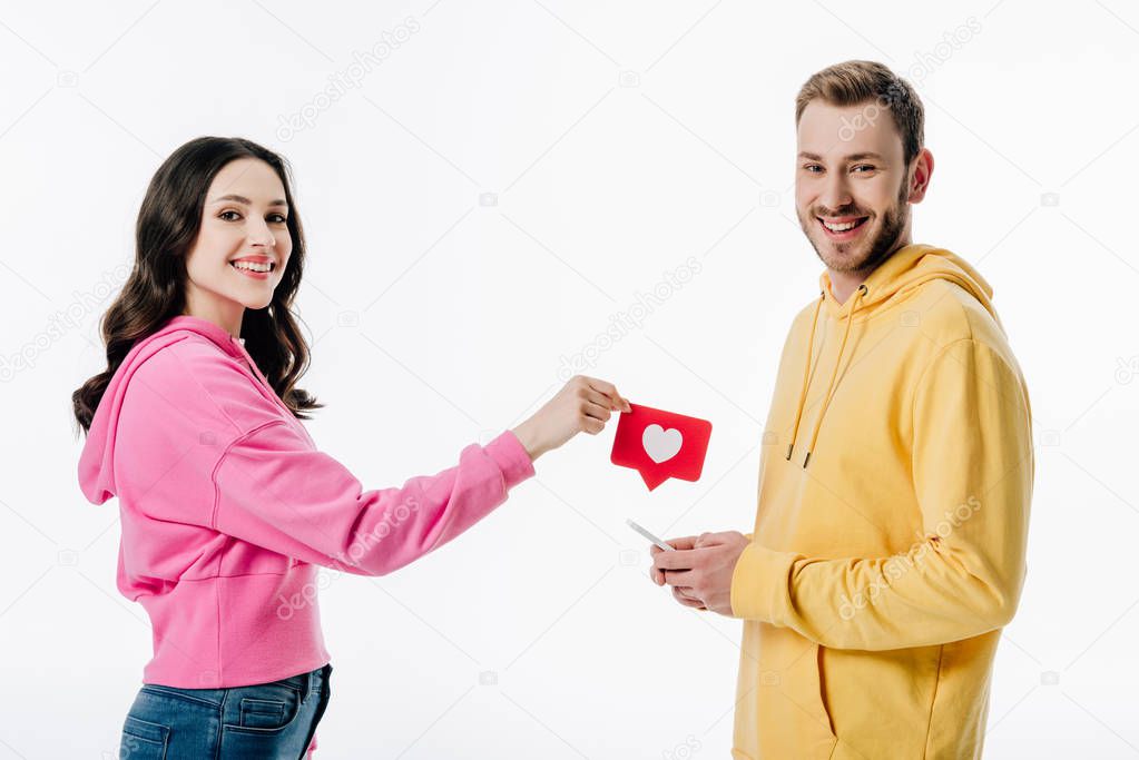 beautiful girl giving red paper cut card with heart symbol to smiling boyfriend with smartphone isolated on white