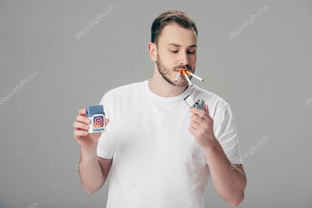 Handsome young man with cigarettes in mouth holding pack with instagram logo isolated on grey