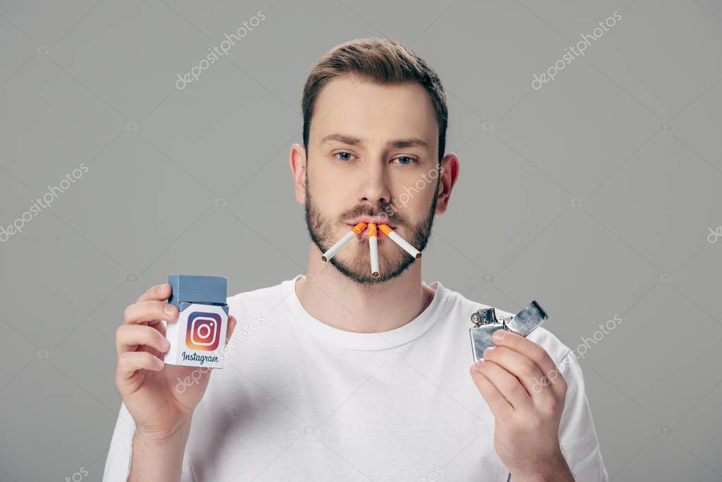 Young man with cigarettes in mouth holding pack with instagram logo and looking at camera isolated on grey