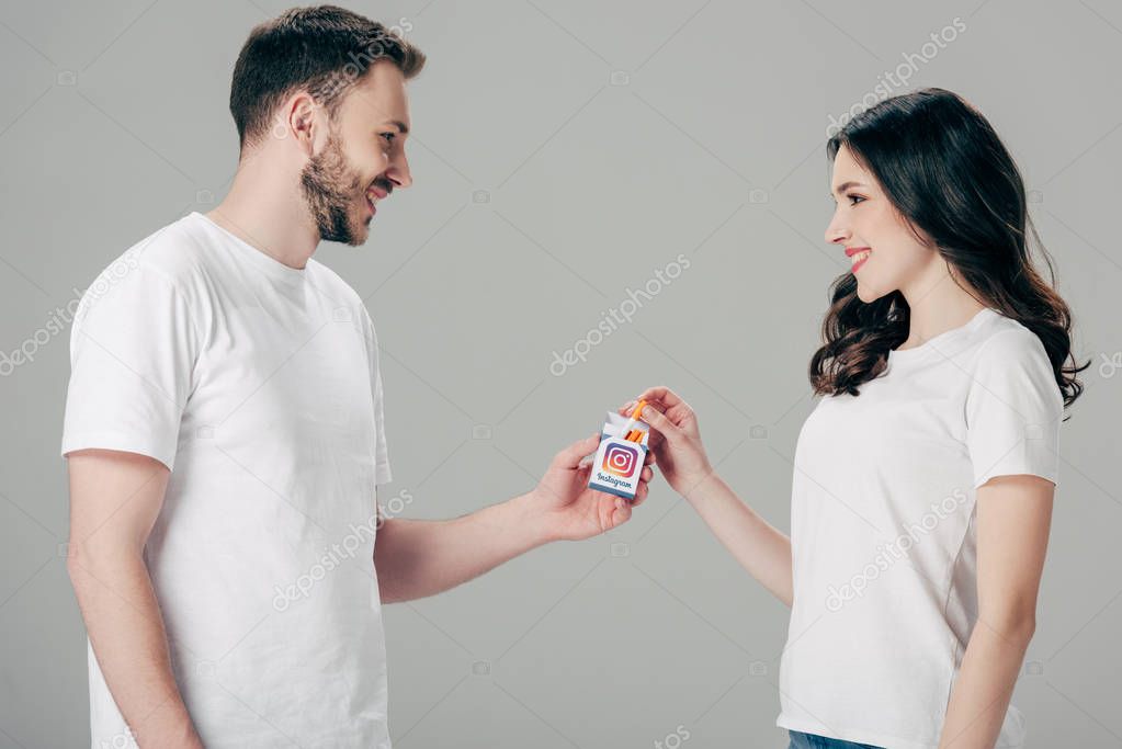 Cheerful man and woman in white t-shirts holding cigarette pack with instagram logo and looking at each other isolated on grey