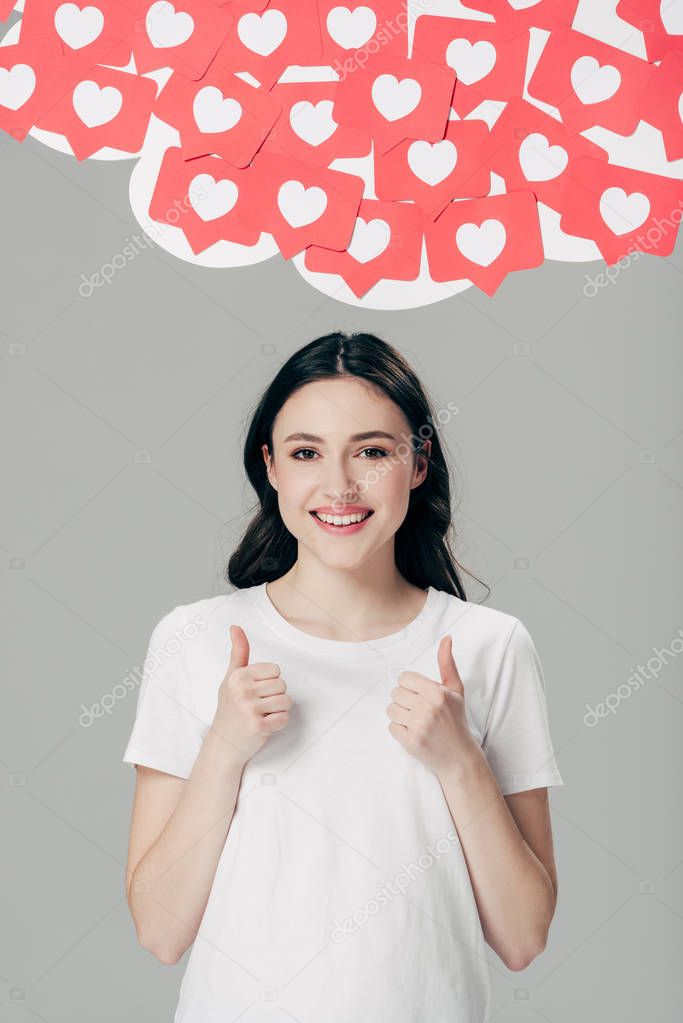 cheerful pretty girl in white t-shirt showing thumbs up near red paper cut cards with hearts symbols isolated on grey