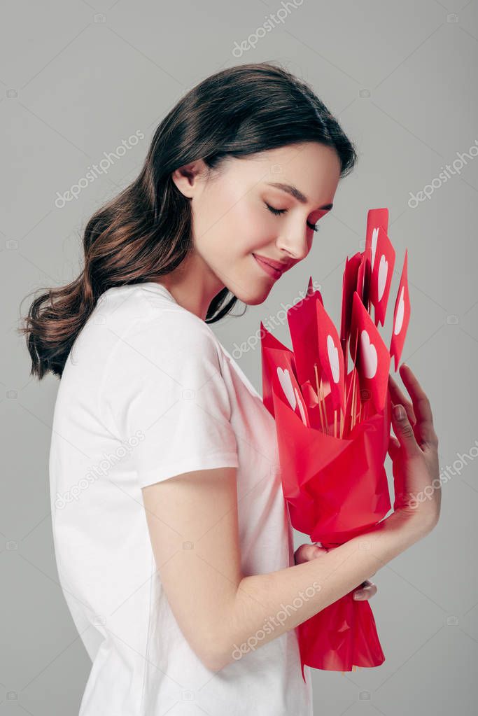 happy dreamy girl holding bouquet of red paper cut cards with hearts symbols isolated on grey
