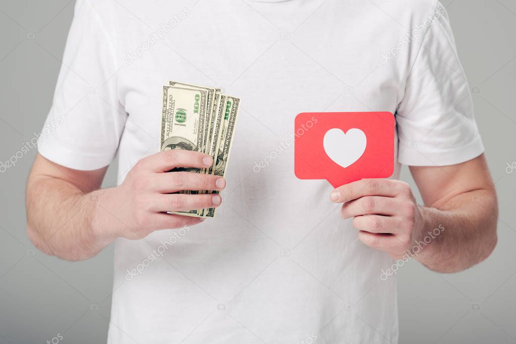 partial view of man holding dollar banknotes and red paper cut card with heart symbol isolated on grey