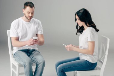 beyaz t-shirt ve mavi kot sandalyeüzerinde oturan ve gri arka plan üzerinde akıllı telefonlar kullanarak erkek ve kadın odaklı
