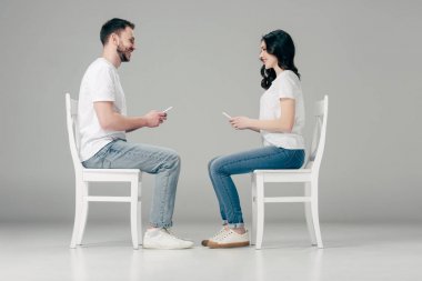 beyaz t-shirt ve mavi kot akıllı telefonlar ile sandalyelerde oturan ve gri arka planda birbirlerine bakarak gülümseyen erkek ve kadın yan görünümü