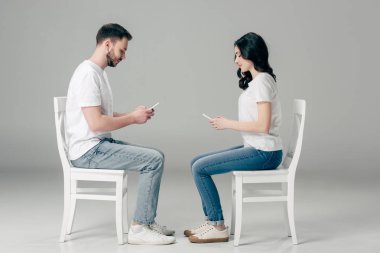 gri arka plan üzerinde sandalyelerde otururken akıllı telefonlar kullanarak beyaz t-shirt ve mavi kot konsantre erkek ve kadın yan görünümü 