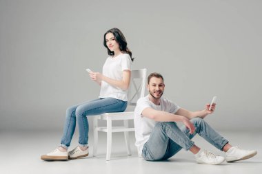 gri arka planda akıllı telefon ile katta oturan neşeli adam yakınında akıllı telefon kullanarak gülümseyen kadın