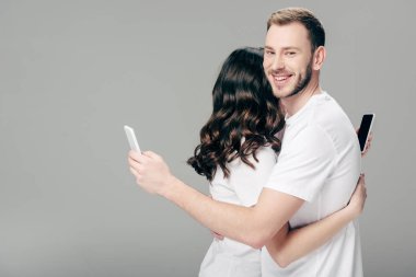 Beyaz tişörtlü genç çift gri arka planda akıllı telefonları kullanırken sarılıyorlar.