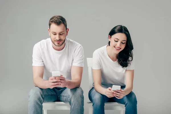 穿着白色T恤和蓝色牛仔裤的年轻夫妇坐在椅子上 用灰色背景的智能手机 — 图库照片