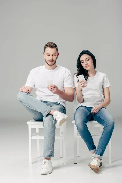 白いTシャツと青いジーンズを着た男女が椅子に座り 灰色の背景にスマートフォンを使用 — ストック写真
