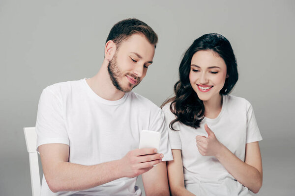 smiling girl showing thumb up while sitting near boyfriend with smartphone on grey background