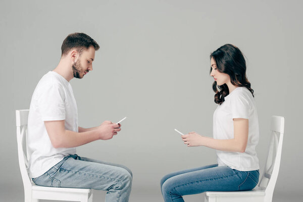side view of focused man and woman in white t-shirts and blue jeans using smartphones while sitting on chairs on grey background