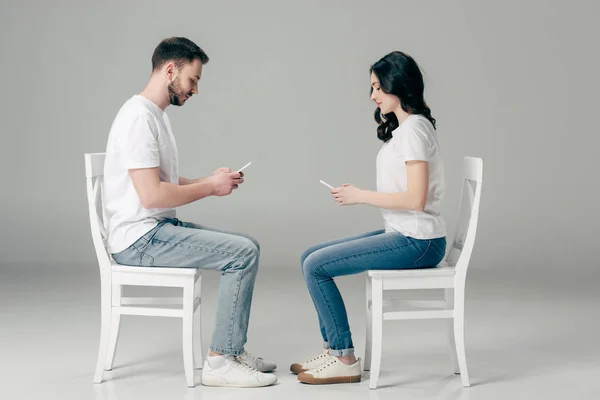 배경의 의자에 앉아있는 스마트 사용하여 티셔츠와 청바지에 남자와 여자의 측면보기 — 스톡 사진