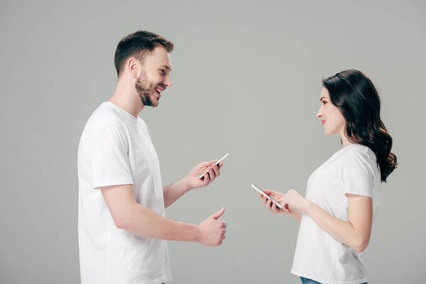handsome man showing thumb up while standing near pretty girl with smartphone isolated on grey