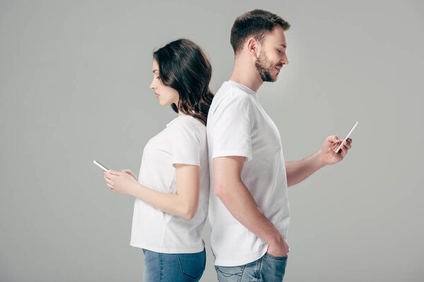 focused man and woman in white t-shirts standing back to back and using smartphones isolated on grey