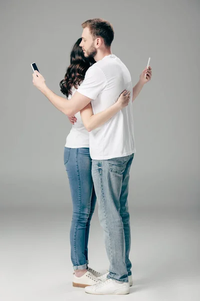 Young Couple White Shirts Blue Jeans Embracing While Using Smartphones — 스톡 사진