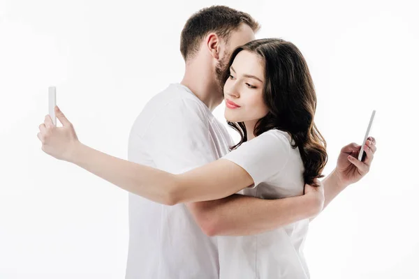 young couple in white t-shirts hugging while using smartphones isolated on white