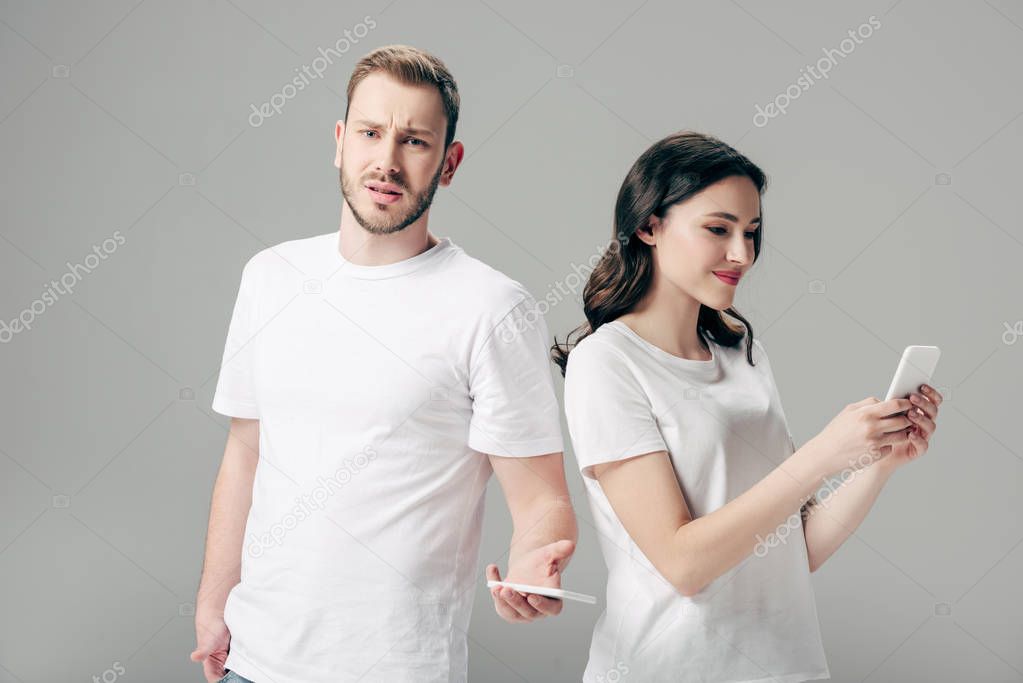 pretty young woman using smartphone near offended boyfriend looking at camera isolated on grey