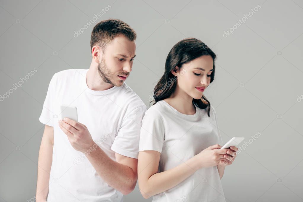 offended young man looking at pretty girlfriend using smartphone isolated on grey