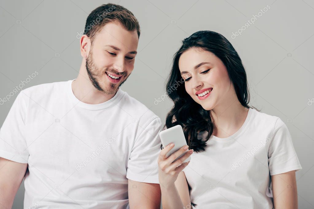 cheerful young man and woman in white t-shirts using smartphone isolated on grey