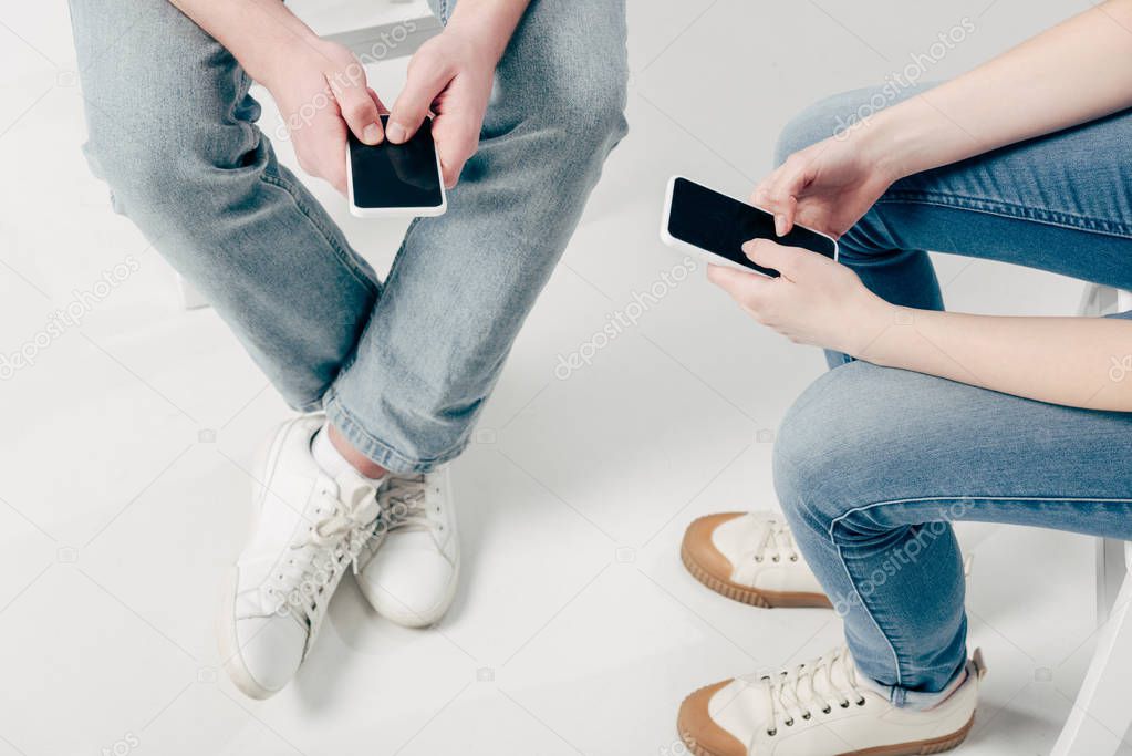 cropped view of man and woman in blue jeans and sneakers using smartphones on white background