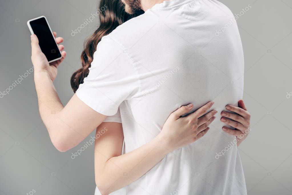 cropped view of woman embracing man using smartphone on grey background