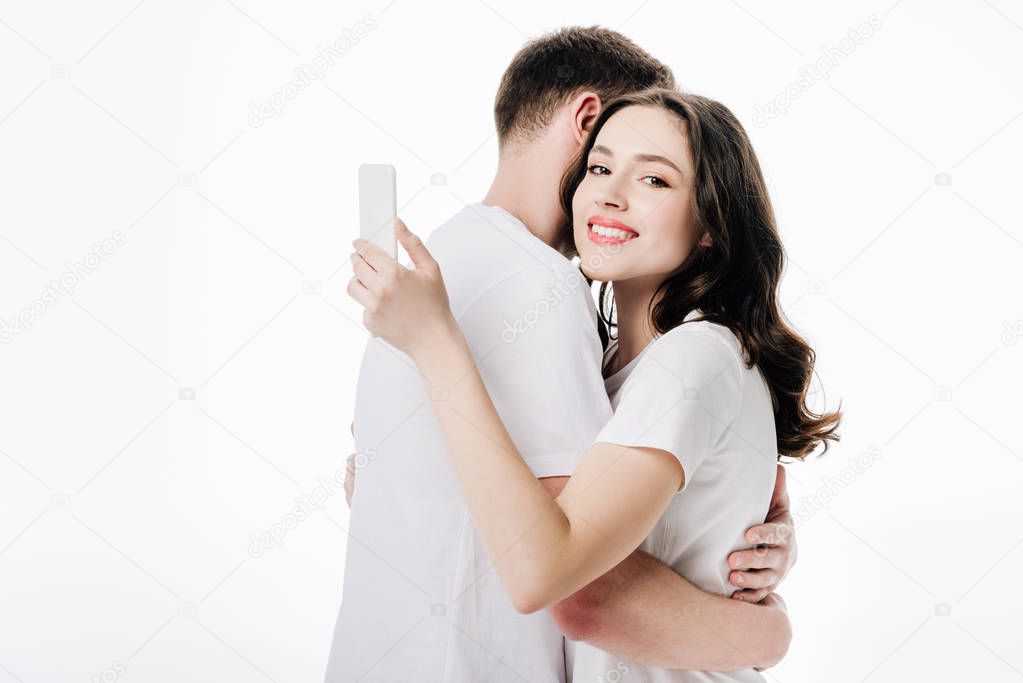 young man hugging pretty girlfriend using smartphone and looking at camera isolated on white