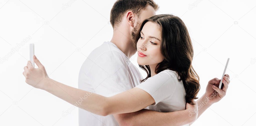 panoramic shot of young couple embracing while using smartphones isolated on white
