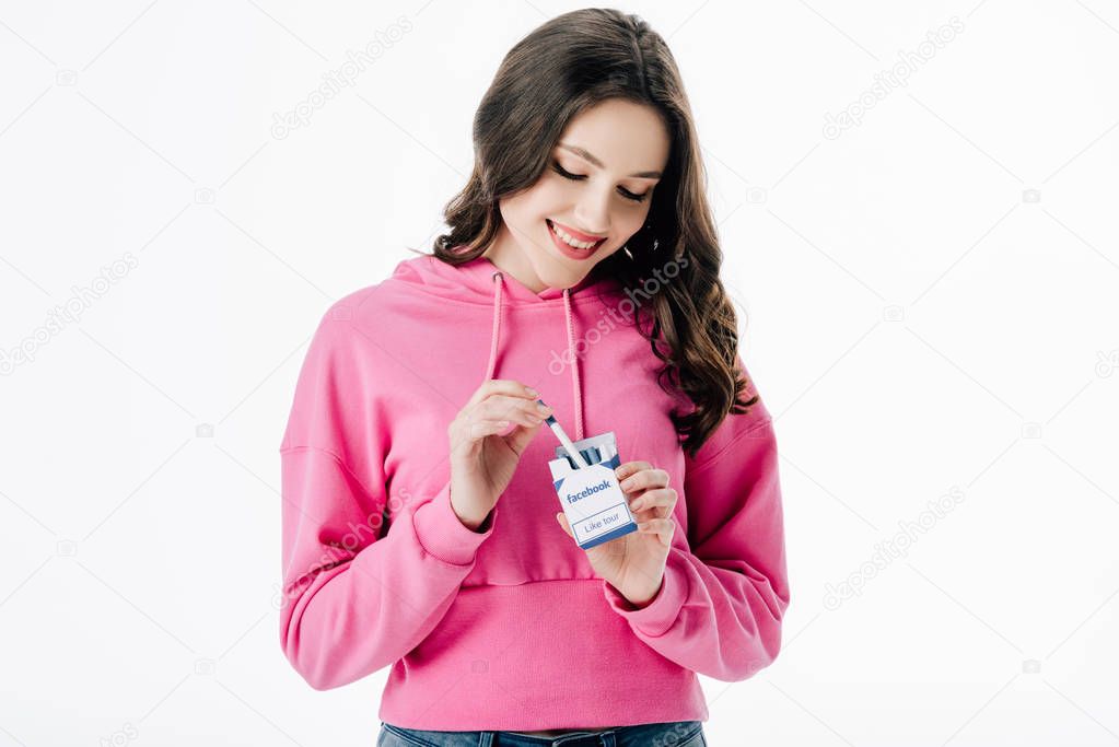 Smiling young woman in pink hoodie getting cigarette from pack with facebook logo isolated on white