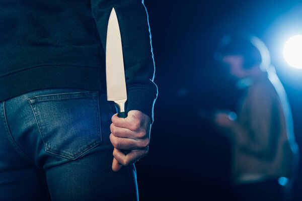 cropped view of murderer hiding knife behind back on black