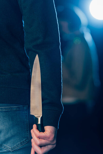 cropped view of murderer hiding knife behind back on black