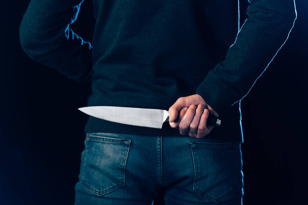 cropped view of murderer hiding knife behind back on black