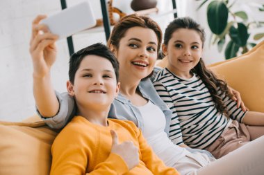 mutlu çekici kadın gülümseyen kızı ve oğlu yakınında kanepede otururken akıllı telefon ile selfie alarak 