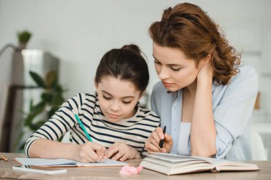 attractive woman helping concentrated daughter doing schoolwork at home clipart
